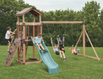 Children playing on wooden backyard swing set with slide, climbing wall, and multiple swings in grassy yard