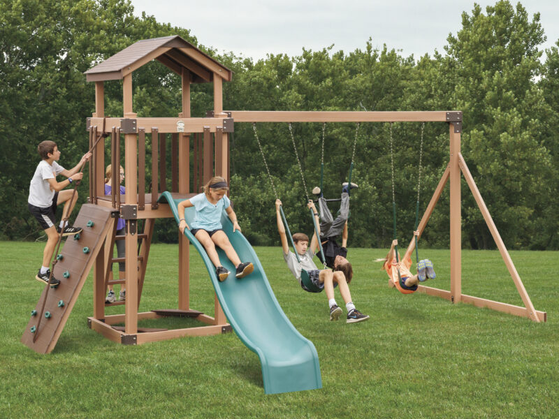 Children playing on wooden backyard swing set with slide, climbing wall, and multiple swings in grassy yard