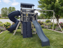 A backyard vinyl swing set including a black tube slide, a climbing wall, and a swing beam, set on a green lawn with flower beds.