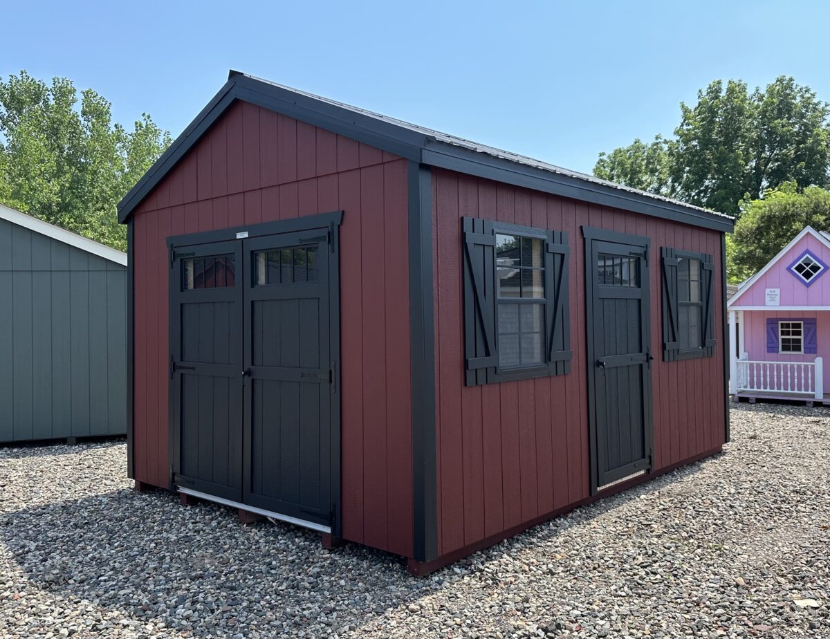 Red and black 12x16 New England style shed with white trim, transom windows in the doors, and a black metal roof on a gravel display lot.