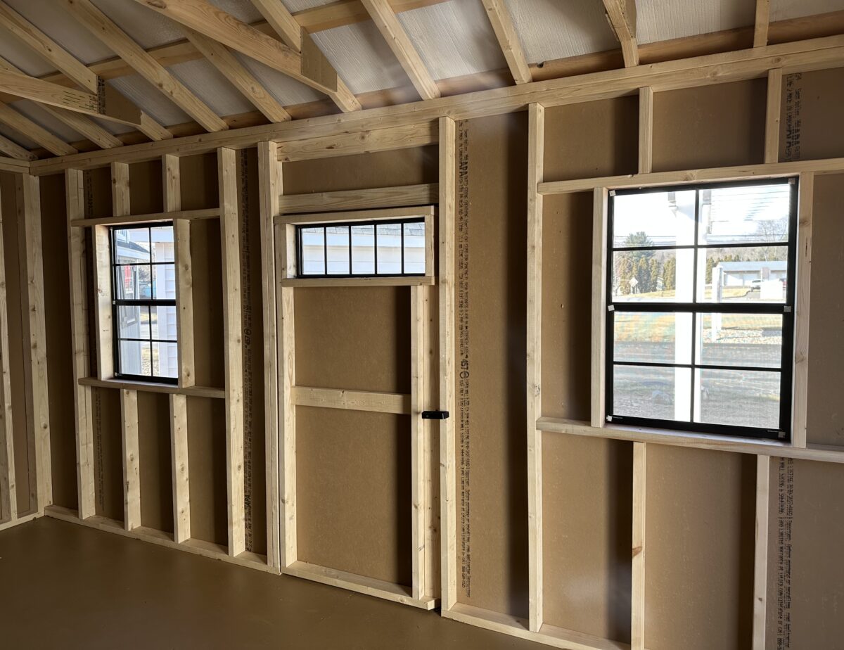 Interior of a 12x16 wood shed showing wooden wall studs, a single entry door with transom windows, and built-in shelving.