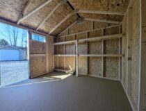 Interior of a 10×10 Madison Peak shed with exposed wood framing, built-in shelving along the side wall, durable flooring, and a transom window above the open double doors providing natural light.