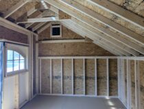 Interior of a 10×16 Carriage House shed with exposed wood framing, open floor space, and a gable vent, with natural light entering through the door window.