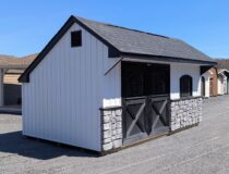 10×16 Carriage House shed with white board and batten vinyl siding, black trim, and rustic black shingles, featuring decorative double doors with arched windows and a stone wainscot accent.