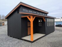 12×20 Offset Studio shed with black siding, pecan trim, and a black textured metal roof, featuring a covered front porch with exposed wood posts, transom windows, and a side wall of multi-pane windows.