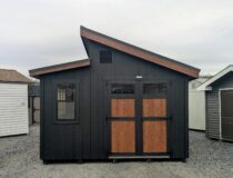 Front view of a 12×20 Offset Studio shed with black siding, pecan trim, and a black textured metal roof, showcasing double doors with transom windows, decorative hardware, and a single window for added natural light