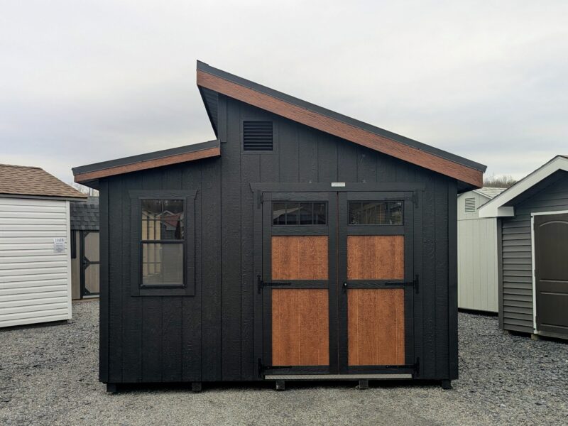 Front view of a 12×20 Offset Studio shed with black siding, pecan trim, and a black textured metal roof, showcasing double doors with transom windows, decorative hardware, and a single window for added natural light