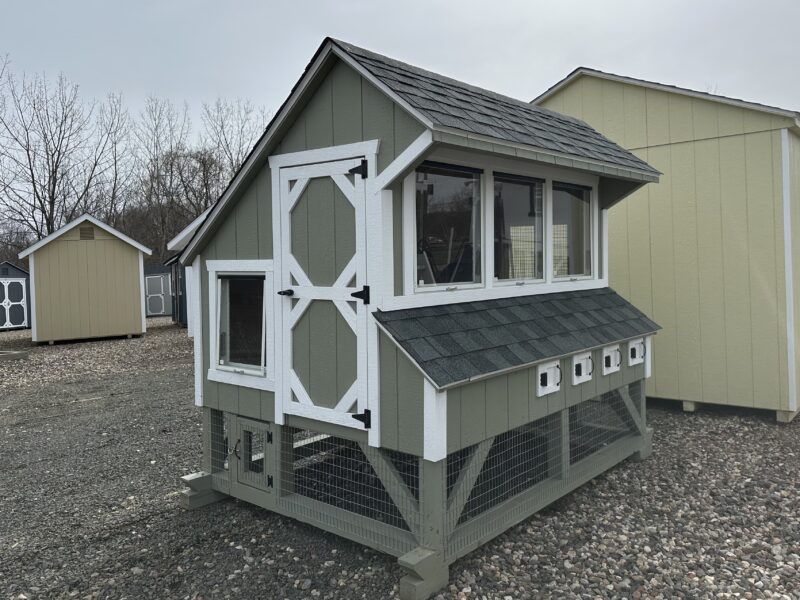 A 6x8 wooden chicken coop with green LP siding, white trim, and a grey shingled roof, featuring a human-sized entry door and windows.