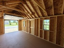 Interior view of an unfinished 10x20 wood storage shed showing the exposed wall framing, roof rafters, windows, and an open overhead garage door.
