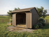 A brown wooden run-in horse shelter with a natural finish, placed in a grassy meadow with a wooden fence in the background.