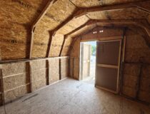 Interior view of an unfinished wooden storage shed with a gambrel roof truss system, OSB wall panels, plywood flooring, and an open double door leading outside.