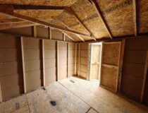 Interior view of an unfinished wooden storage shed with a peak roof truss system, OSB wall panels, plywood flooring, and an open double door leading outside.