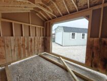 The interior perspective looking out from a wooden run-in shelter, showing wall studs, wood paneling, and a gravel floor.