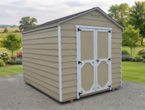 Angled view of a tan wooden storage shed with white trim and double doors, surrounded by a scenic backyard with green grass and trees.