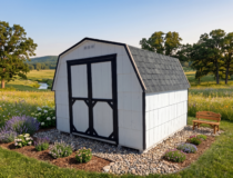 A white wood-sided storage shed with a black shingled gambrel roof, black trim, and double barn-style doors, sitting on a gravel lot next to other outdoor structures.